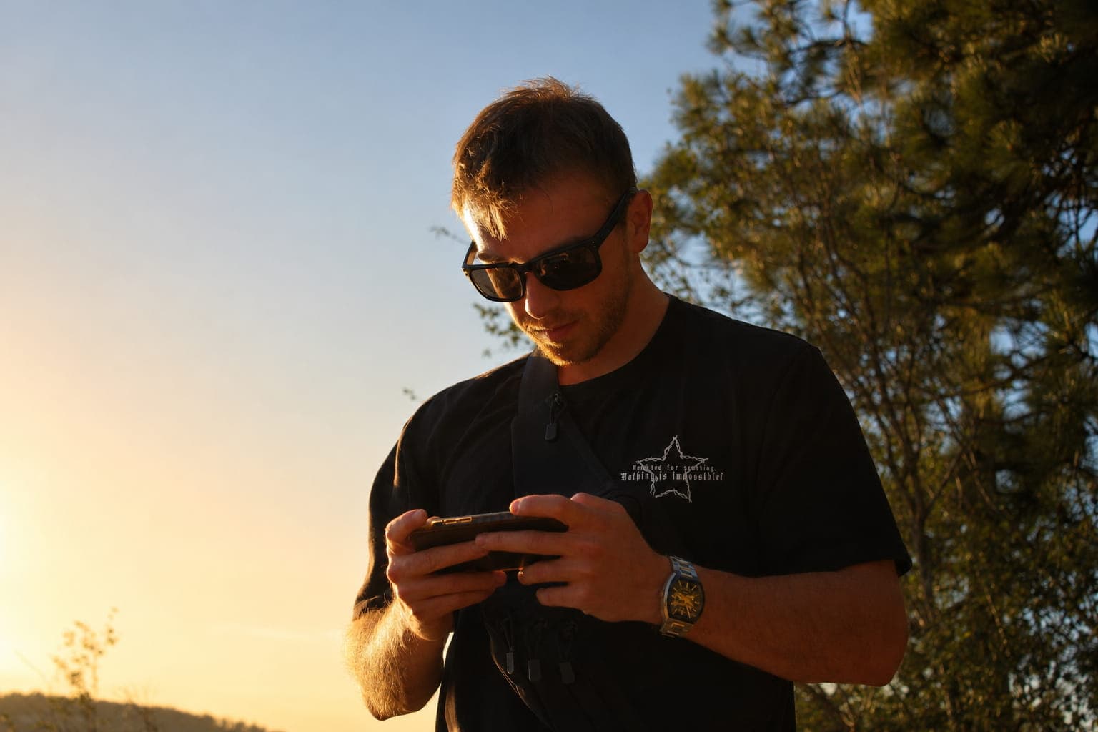 Man wearing sunglasses looks at his smartphone during golden hour with trees in the background.