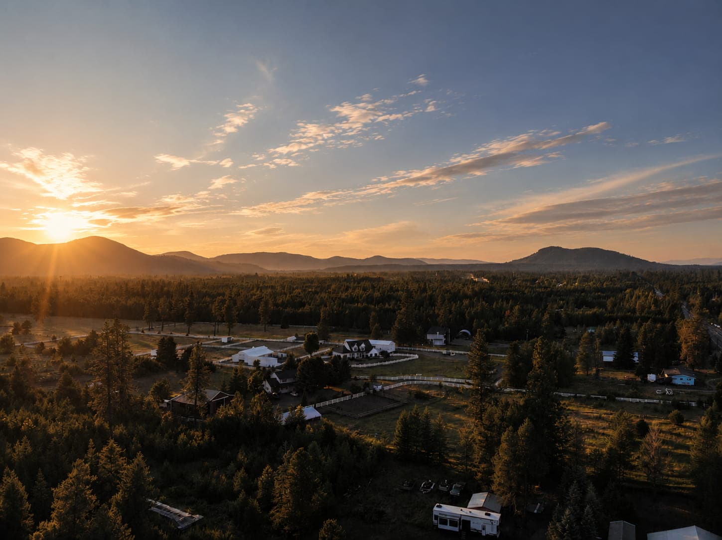 Golden sunset over a forested valley with rural farmhouses and mountains in the distance.