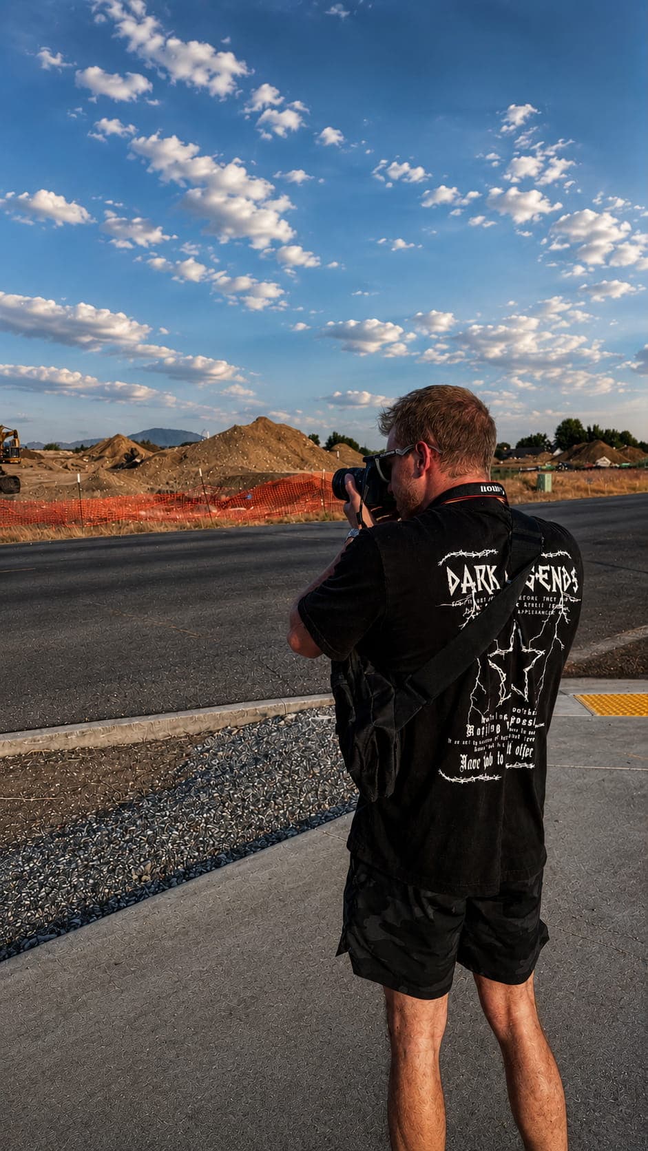 Man photographing dirt mounds at a construction site under a blue sky with white clouds.