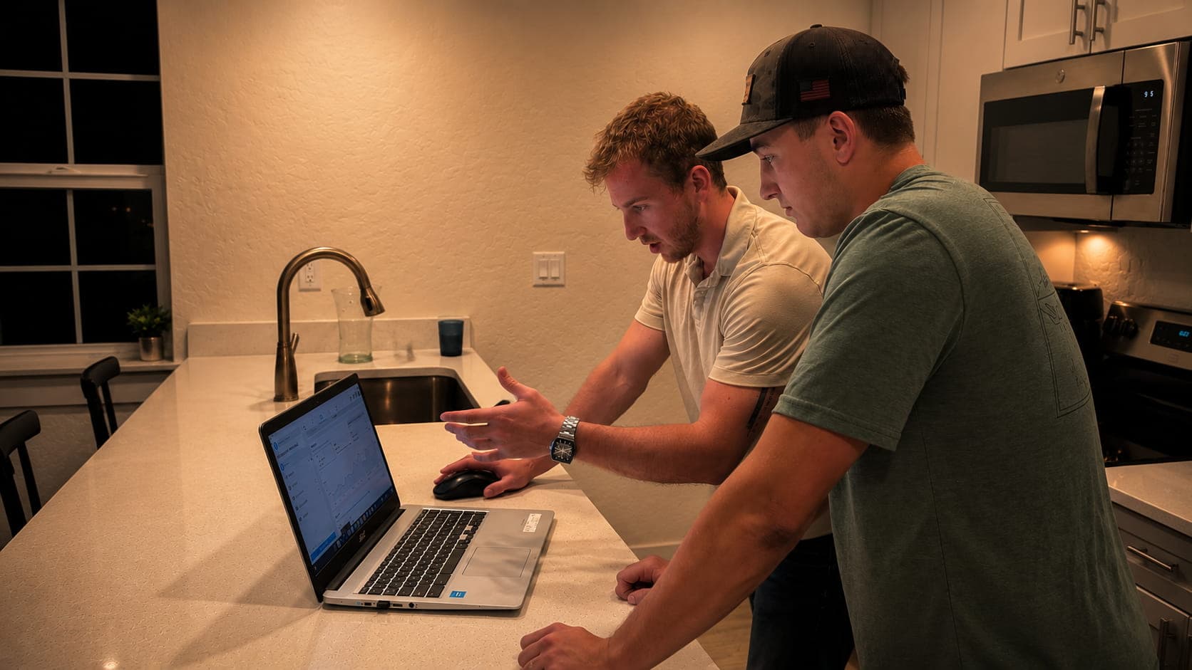 Two men look at a laptop on a kitchen counter, one gesturing at the screen.
